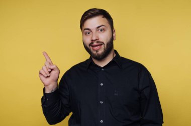 Bir adam sarı arka planda duruyor. Sakalı var ve siyah bir gömlek giyiyor. Yüzünde neşeli bir ifadeyle yukarıya doğru işaret ediyor. Bir fikri ya da düşüncesi olduğunu gösteriyor..