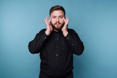 Stüdyoda mavi bir geçmişi olan bir adam var. İki eliyle yüzüne dokunuyor. Siyah bir gömlek giyerken bir şey düşünüyor ya da düşünüyor gibi görünüyor..