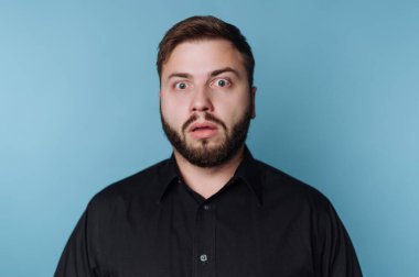 Açık mavi bir duvara karşı duran bir adam, ağzı ve gözleri açık şaşırmış bir yüz ifadesi sergiliyor. Şoka girmiş ya da durumu kabullenemiyor gibi..