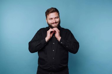 Sakallı bir adam mavi bir arka plana karşı duruyor. Gülümsüyor ve ellerini yüzünün yanında tutuyor. Siyah gömleği basit bir ayara ekleniyor..