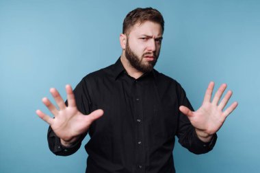 Bir adam elleri havada mavi bir zemine karşı duruyor. Ciddi görünüyor ve sahnede görünmeyen bir şeye reaksiyon gösteriyor. İfadesi emin olmadığını gösteriyor..