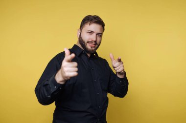 Bir adam sarı bir arka planın önünde duruyor, gülümsüyor ve parmaklarını kameraya şakacı bir şekilde doğrultuyor. Siyah bir gömlek giyiyor ve kendinden emin görünüyor..