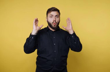 Adam elleri havada ve ağzı açık duruyor, şaşırıyor. Siyah bir gömlek giyer ve stüdyo seansı sırasında sarı bir arkaplan önünde poz verir..