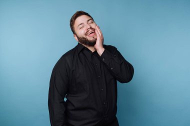 Bir adam mavi bir arkaplanın önünde duruyor. Yüzüne tek eliyle dokunarak geniş bir gülümseme sergiliyor. Gömleği koyu renk. Bu sahne stüdyo ortamında geçiyor..