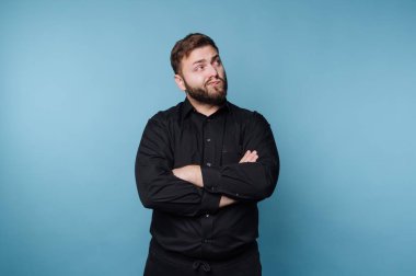 Kollarını kavuşturup düşünceli bir ifadeyle yukarı bakan bir adam. Siyah bir gömlek giyiyor ve mavi arka planı olan bir stüdyoda. Ayar gündelik ve parlak görünüyor..