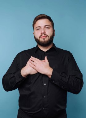 Mavi arka planı olan bir adam içeride duruyor. Siyah bir gömlek giyiyor ve iki elini de göğsüne koyuyor. Ayarlar basit ve ışıklandırması iyi..