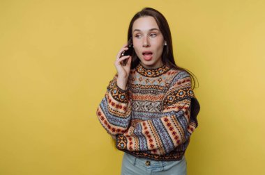 Bir kadın, kulağına telefon dayayan parlak sarı bir arka plana karşı duruyor. Renkli bir kazak giyerken şaşırmış görünüyor. İfadesi çağrıdaki bağlılığı gösteriyor..