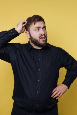 A man with a beard stands against a bright yellow background. He has a confused look on his face and has one hand on his hip and the other scratching his head.