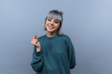 Genç bir kadın gri bir fonda duruyor ve parmaklarıyla el hareketi yaparken gülümsüyor. Kısa saçları var ve bol bir kazak giyiyor..