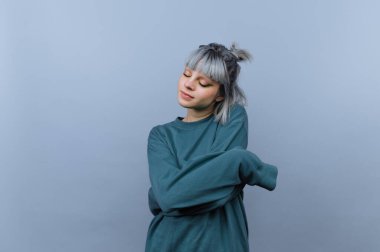 Genç bir kadın kollarını düz mavi bir arkaplanda kavuşturmuş duruyor. Açık gri saçları var ve uzun kollu bir gömlek giyiyor. Işıklar yumuşak ve eşit..