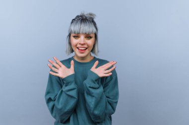 Kısa gri saçlı bir kişi açık mavi arka planı olan kapalı bir yerde duruyor. Coşku ve neşe göstermek için ellerini kaldırırken geniş bir gülümseme sergiliyorlar. Giysileri rahat ve rahat..