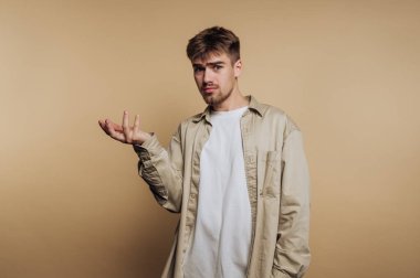 Genç bir adam bej bir arka plana karşı sorgulayıcı bir bakışla duruyor. Kısa saçlı ve beyaz bir tişörtün üzerine açık renk bir gömlek giyiyor. El hareketleri kafa karışıklığını ya da soruşturmayı gösteriyor..