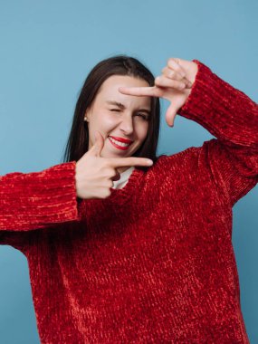 Mavi bir duvarın önünde genç bir kadın duruyor. Gülümser ve ellerini çerçeve oluşturmak için kullanır. Onun kırmızı kazağı sette göze çarpıyor. Atmosfer dost canlısı ve rahat..