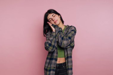 Genç bir kadın pembe bir duvara yaslanmış, üzerinde yeşil bir tişört var. Gözlerini kapar ve ellerini yüzüne tutar. Huzurlu bir andan zevk alıyormuş gibi görünür..