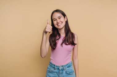 Genç bir kadın düz sarı bir duvarın önünde duruyor. Uzun siyah saçları var ve gülümsüyor. Sağ eliyle baş parmağını kaldırarak hafif bir anın tadını çıkarıyor..