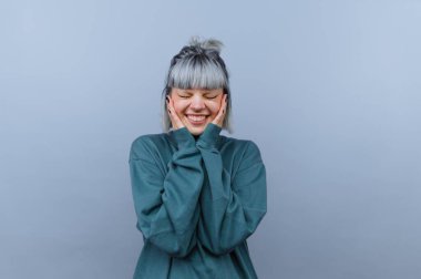 Gümüş saçlı genç bir kadın gülümseyerek ve elleriyle yüzünü örterek mutluluğu gösterir. Düz bir arkaplanda duruyor..