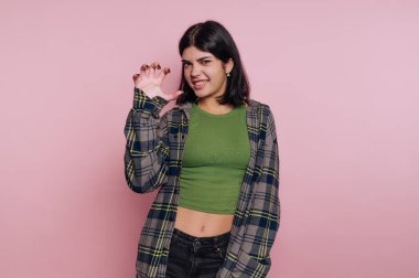 Pembe bir duvarın önünde duran kişi neşeli bir el hareketi yapıyor. Yeşil bir bluz ve ekose gömlek giyiyorlar. Ayarlar basit ve onların ifadesine odaklı..