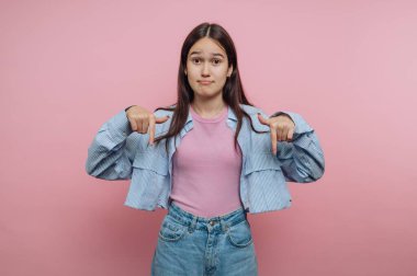 Genç bir kız pembe bir arka planın önünde duruyor. Kafası karışmış görünüyor ve yere doğru işaret ediyor. Çizgili bir ceket ve kot pantolonlu açık renk bir gömlek giyiyor..