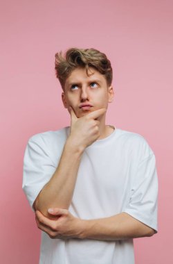 Genç bir adam tek eliyle çenesinde duruyor, düşünürken yukarı bakıyor. O, katı pembe bir arka planın önünde. Odadaki ışık parlak ve berrak..