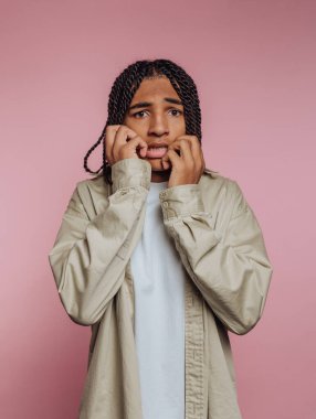 Genç bir insan pembe bir duvarın önünde endişeli görünüyor. Elleri ağızlarına yakın, endişe belirtileri gösteriyor. Stüdyo ayarı net ve odaklanmış bir görünüm veriyor..