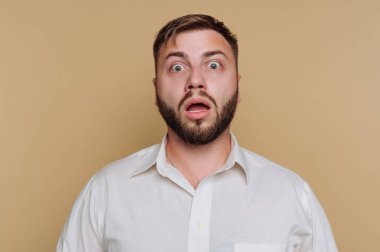 Kahverengi bir arka planın önünde şaşırmış bir ifadeyle duran bir adam. Gözleri kocaman ve ağzı açık, bu da şok ya da inançsızlığı gösteriyor. Beyaz bir gömlek giyiyor..