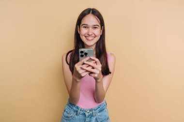 Genç bir kadın gülümseyerek ve akıllı telefonunu tutarak düz bir arkaplanın önünde duruyor. Mutlu ve nişanlı görünüyor. Sahne kapalı bir yerde geçiyor..