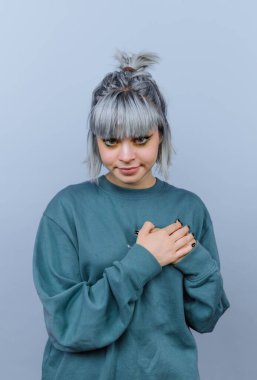 Bir kişi açık mavi bir arkaplanın önünde duruyor. Gri saç şekilleri sıradan ve yeşil bir kazak giyiyorlar..