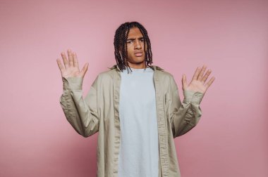 Genç bir adam sorgulama jesti için ellerini kaldırdı. Örgüleri var ve basit bir gömlek giyiyor. Arka plan, gündelik bir ortam gibi görünen açık pembe bir renk..