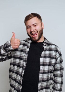 Bir adam arka planda durup gülümserken başparmağını kaldırır. Siyah beyaz kareli bir gömlek giyiyor ve neşeli bir ifade sergiliyor..