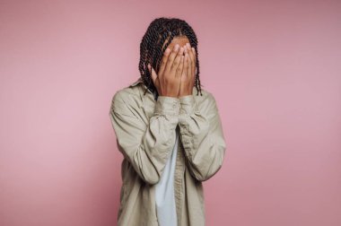 Bir insan sırtını hafifçe dönerek iki eliyle yüzünü kapatarak ayakta durur. Arkaplan koyu pembe bir renktir. Gün ışığında içerisi kapalı gibi görünüyor..