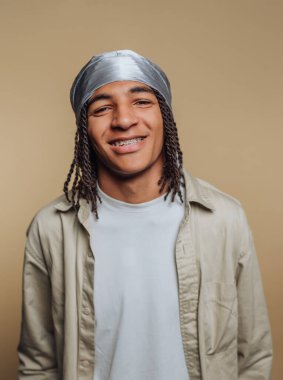 Tarafsız bir arkaplanın önünde duran genç bir adam. O gümüş bir baş örtüsü ve açık renk bir gömlek giyer. Kameraya bakarken gülümsüyor, diş tellerini gösteriyor..