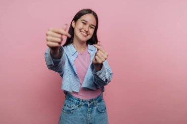 Genç bir kadın pembe bir duvarın önünde duruyor. Gülümsüyor ve parmaklarıyla kalp şekli oluşturuyor. Açık mavi bir ceket ve pembe bir gömlek ve kot şort giyiyor..