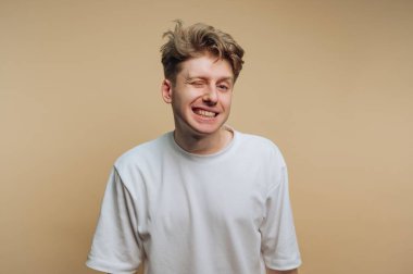 Genç bir adam sade bir arka planın önünde duruyor. Kameraya gülümseyip göz kırpıyor. Onun ifadesi neşeli bir ruh hali gösteriyor. Basit beyaz bir tişört giyiyor..