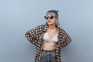 Genç bir kadın açık mavi bir duvarın önünde poz veriyor. Güneş gözlüğü, kareli gömlek ve günlük pantolon giyiyor. Saçları kısa ve boyalı. O, tarzıyla oynarken kendinden emin görünüyor..