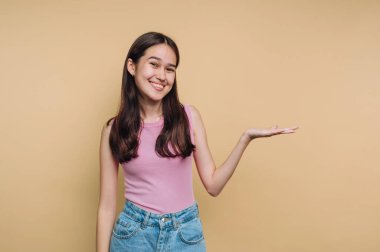 Açık pembe bluzlu ve kot pantolonlu bir kız sarı bir duvara dayanıyor. Gülümser ve elini yana uzatır. Nişanı davet eder..