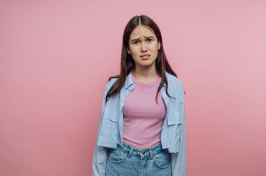 Mavi gömlek ve kot pantolon giyerken kafası karışmış bir kız pembe bir duvara dayanıyor. Uzun saçları var ve kameraya poz verirken şaşırmış görünüyor..