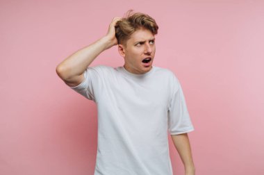 Bir adam pembe bir duvara karşı duruyor, kafası karışmış bir şekilde kafasını kaşıyor. Kafası karışmış gibi görünüyor ve beyaz bir gömlek giyiyor..