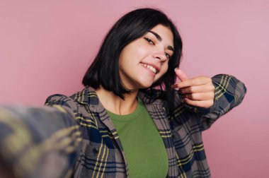 Genç bir insan pembe bir duvarın önünde duruyor, gülümsüyor ve selfie çekerken el hareketi yapıyor. Yeşil bluzun üstüne fanila giyiyorlar..