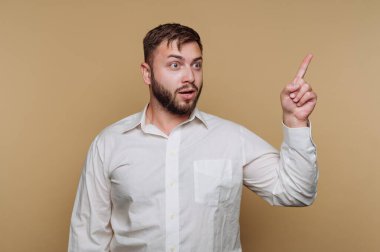 Beyaz gömlek giyen bir adam, yüzünde şaşırmış bir ifadeyle yukarıya doğru işaret eder. Sade bir bej arka planın önünde duruyor. Dekor büyük ihtimalle bir stüdyo..