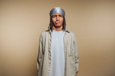 Tarafsız bir ifadeyle duran genç bir adam gümüş bir başörtüsü ve açık renk bir gömlek giyiyor. Arka plan sade ve basittir, kişinin ifadesine odaklanır..