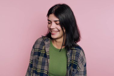 Genç bir kadın pembe bir duvarın önünde duruyor. O, yeşil bir gömlek ve ekose bir gömlek giyer. Onun saçı koyu ve kısa. Gülümsüyor ve rahatlamış görünüyor, anın tadını çıkarıyor..