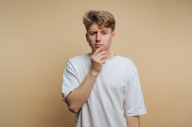 Genç bir adam eli çenesinde, düşünceli ya da meraklı bir şekilde poz verir. Sade bir bej arka planı olan bir stüdyoda duruyor. Yüzü bir şey düşündüğünü gösteriyor..