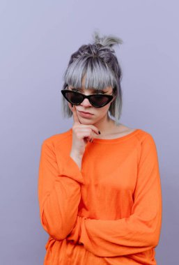 Bir kadın stüdyo ortamında poz veriyor. Mor bir arka planı var. Güneş gözlüğü takıyor ve düşünceli görünüyor. Turuncu bir bluz giyerken çenesini dinlendiriyor..