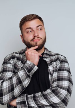 Bir adam kollarını kavuşturarak içeride duruyor. Hafifçe yukarı bakarken yüzünde düşünceli bir ifade var. Siyah bluzunun üstüne ekose gömlek giyer..