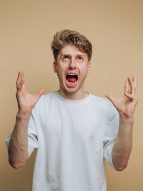 Genç bir adam ellerini başının üstüne kaldırarak ayakta duruyor. Açık bir ağzı var, basit bir arka planda güçlü bir öfke ifadesi gösteriyor..