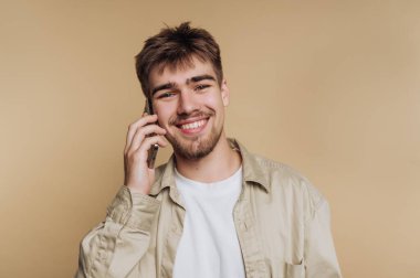 Genç bir adam bir telefon görüşmesi yapıyor. O, düz bir arka planın önünde dururken yüzünde büyük bir gülümseme var. Açık renk bir gömlek giyiyor ve rahat ve mutlu görünüyor..