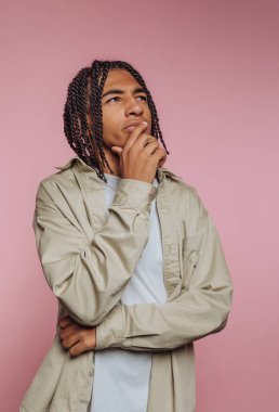 Genç bir adam pembe bir geçmişi olan bir stüdyoda duruyor. O, saçını ördü ve elini çenesinde dinlendirirken düşünceli bir şekilde bakıyor. Açık renk bir gömlek giyiyor ve düşüncelere dalmış gibi görünüyor..