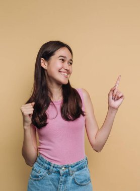 Açık pembe bluzlu ve kot şortlu genç bir kadın ayakta duruyor. O, parlak bir şekilde gülümser ve heyecanlı bir şekilde parmağını havaya kaldırır. Arkaplan sade ve sarı renkte..