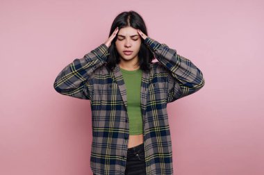 Elleri başının üstünde duran bir kadın rahatsızlık ya da stres gösterir. O, ekose bir gömleğin altına yeşil bir üst giyer. Arka plan pembe ve gündüz..