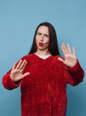 Kırmızı bir kazak giyen bir kadın mavi bir arka fonun önünde duruyor. El hareketleri yapıyor ve yüzünde şakacı bir ifade var. Stüdyo onun hareketlerine odaklanıyor..